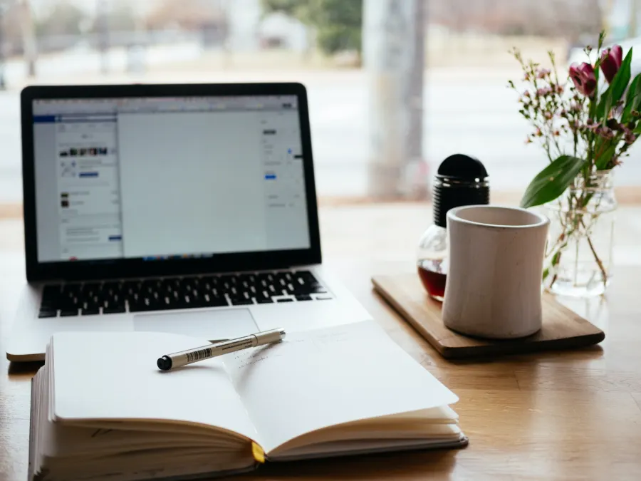 A laptop and an open blank notebook with a pen, beside a coffee mug and flowers on a desk by a window.